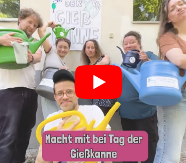 Eine Gruppe von Menschen mit Gießkannen stehen vor einem Plakat "Tag der Gießkanne 4. Juli". Vorn im Bild steht der Schriftzug "macht mit beim Tag der Gießkanne"
